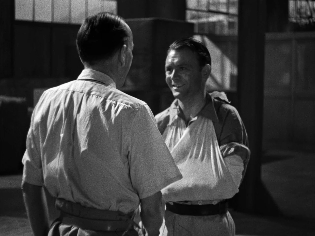 In Which We Serve (1942) movie still: medium — An industrial building with large metal-framed windows and stacked crates in the background. A man…