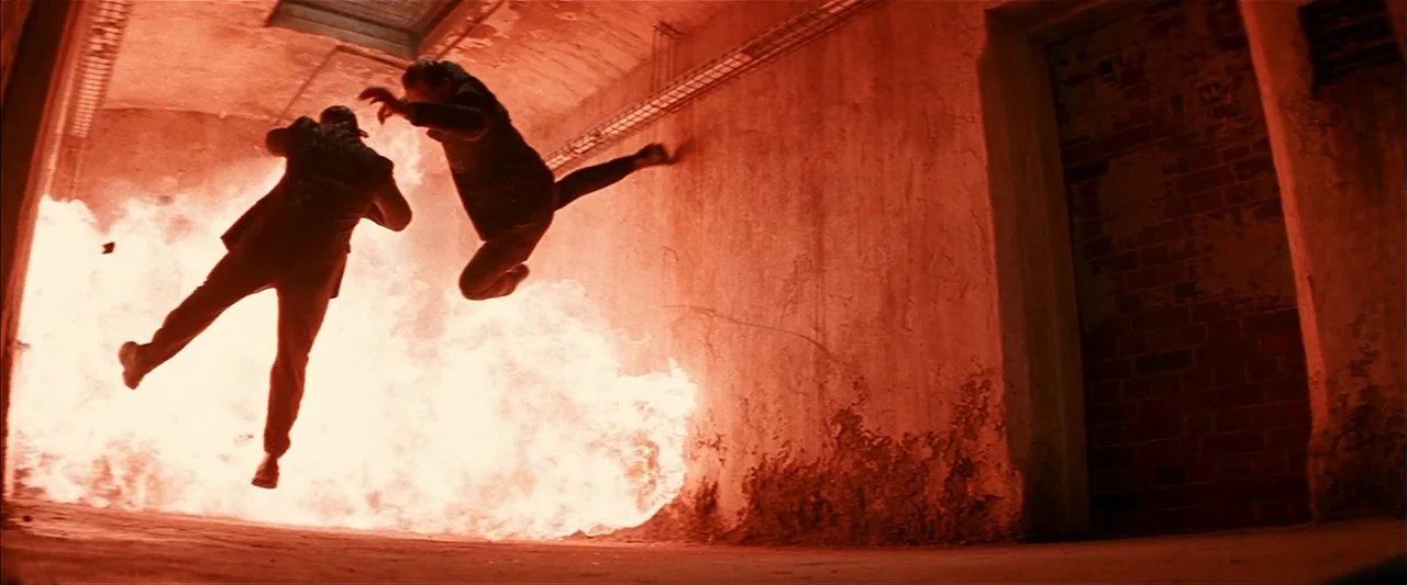 Mission: Impossible II (2000) movie still: wide — An industrial concrete passage with brick walls is engulfed by a violent, bright explosion. Two…