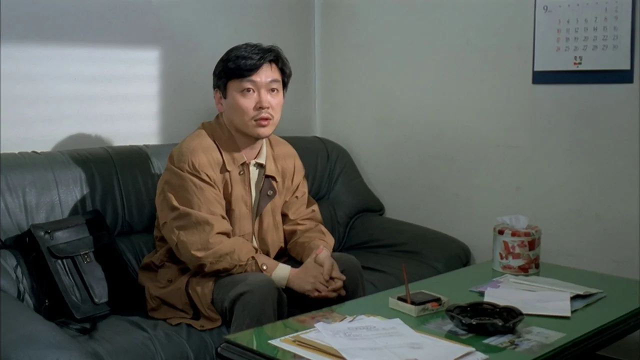 The Day a Pig Fell Into the Well (1996) movie still: medium — A man sits on a dark leather couch in a plain-walled office space. Papers and a tissue…