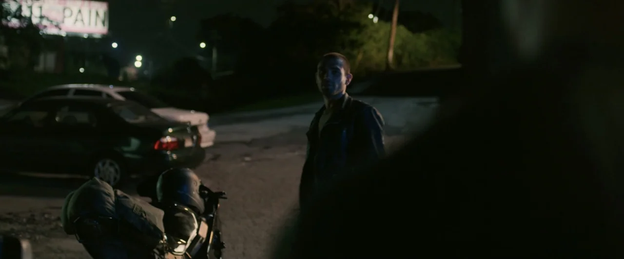 One Fast Move (2024) movie still: medium — A dark outdoor parking lot at night with parked vehicles and distant illuminated signs. A man in a dark…
