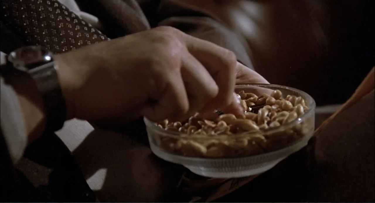 Gorky Park (1983) movie still: closeup — A glass bowl filled with salted peanuts rests on a dark surface. A hand wearing a silver wristwatch reaches…
