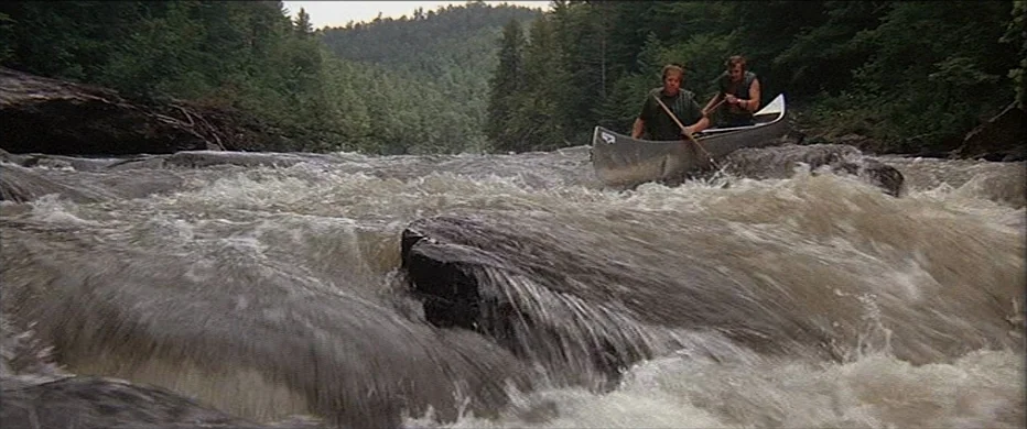 Deliverance (1972) movie still: wide — A wide river with fast-moving, turbulent water flows through a dense wooded valley. Two men in a silver canoe…