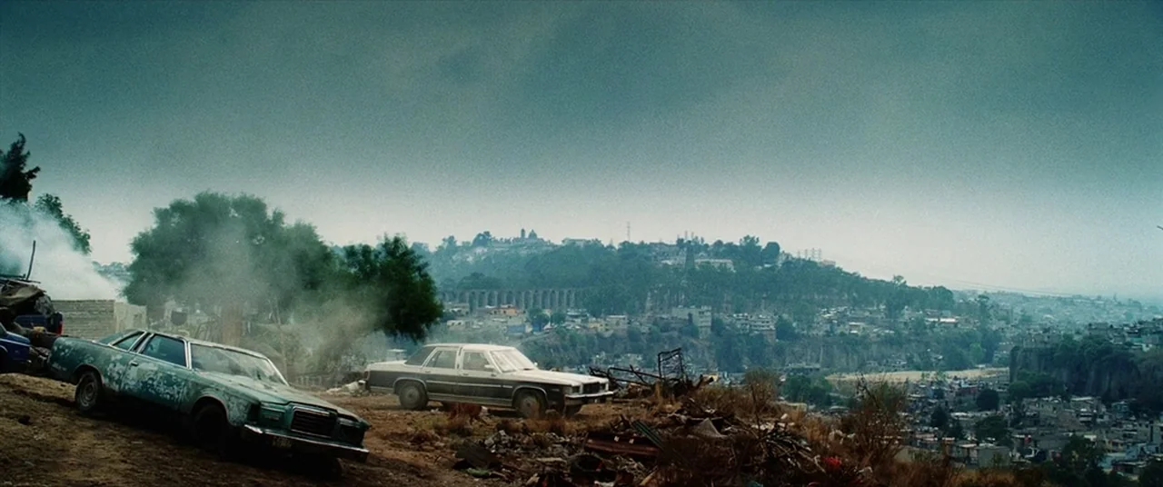Man on Fire (2004) movie still: wide — A hillside junkyard contains two abandoned sedans positioned among scattered debris and dry grass. The…