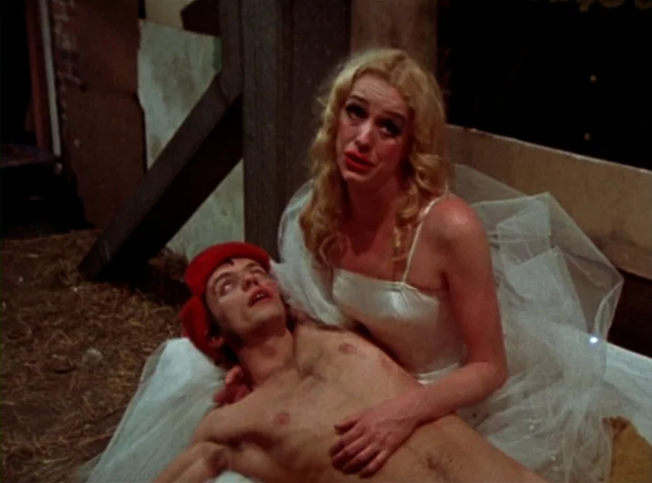 Desperate Living (1977) movie still: medium — A performance area covered in scattered straw with wooden beams visible in the corner. A woman kneels…