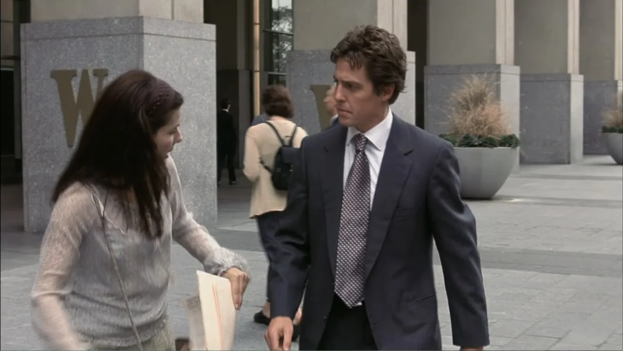 Two Weeks Notice (2002) movie still: medium — A stone-paved plaza outside a modern office building with large grey concrete pillars. A woman in a…