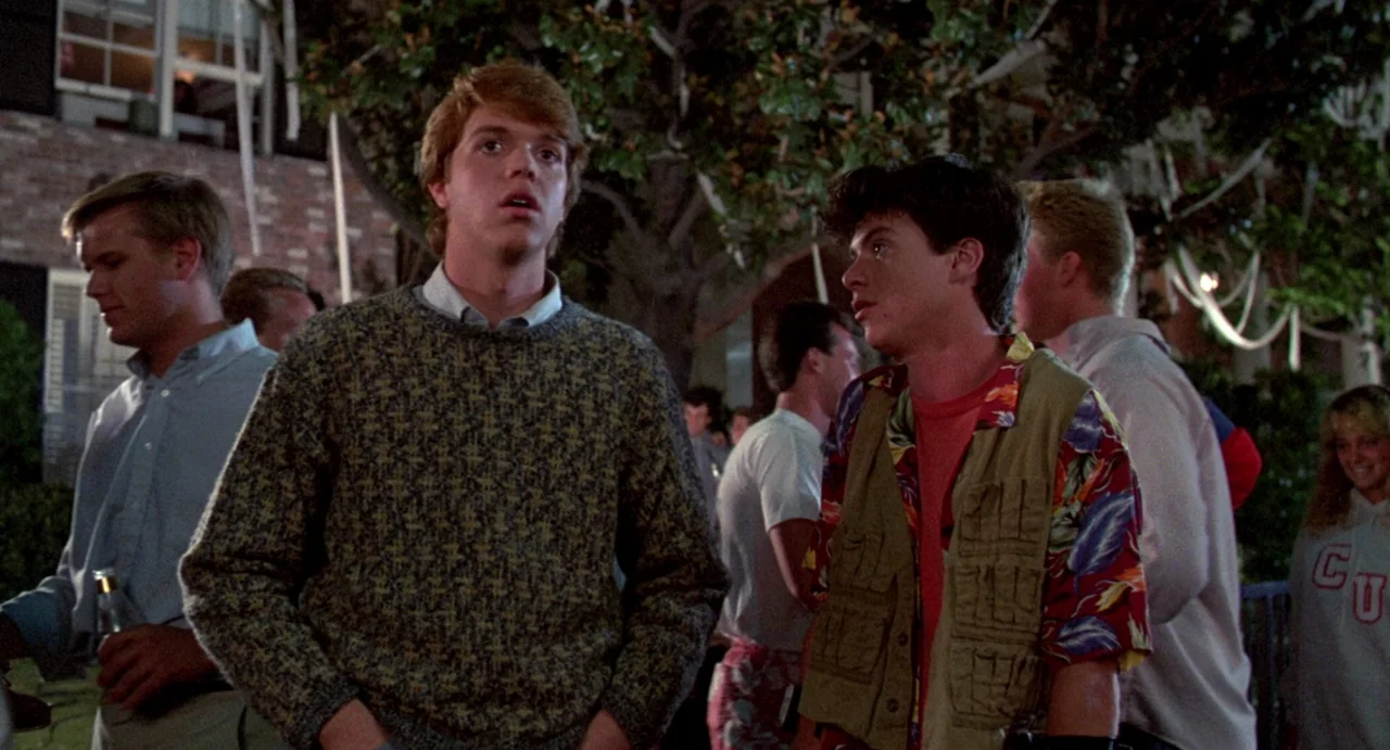 Night of the Creeps (1986) movie still: medium — A dark outdoor space with trees decorated with hanging streamers in the background. Two young men…