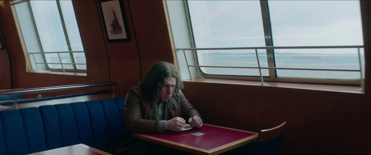 Border movie still: medium — A quiet passenger area on a ferry with blue bench seating and wood walls. A man sits at a small table near a window,…