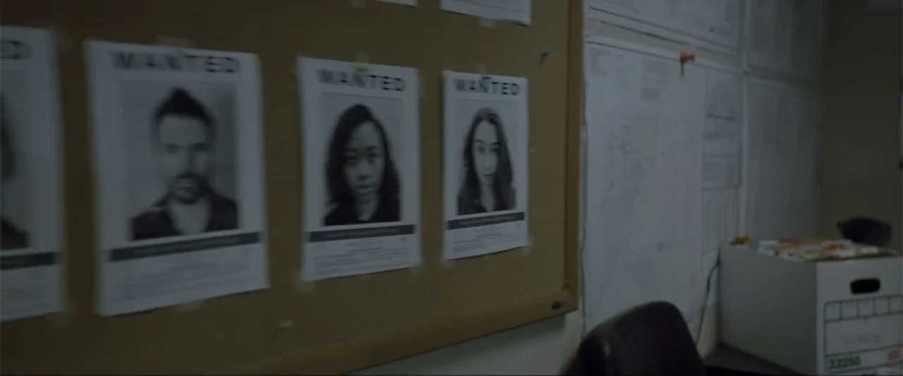 Gender Under Attack (2018) movie still: medium — A corkboard holds several wanted posters with photographs of people in an office. A storage box…
