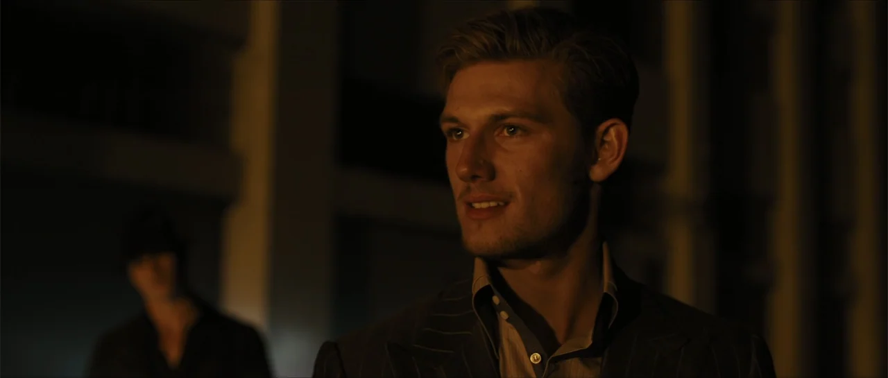 In Time (2011) movie still: medium — A dimly lit hallway features a man in the foreground and a blurred figure behind him. The man is wearing a…