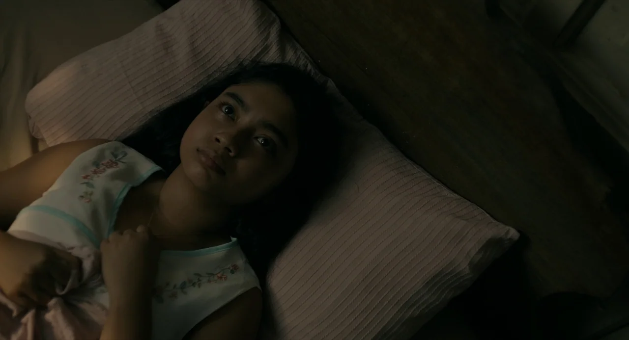 In My Mother’s Skin (2023) movie still: closeup — A person lies on a textured pillow in a dark, dimly lit bedroom. The individual gazes upward while…
