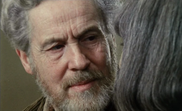 After The Rehearsal (1984) movie still: closeup — An interior space with a plain, neutral-colored wall in the background. An elderly man with a grey…