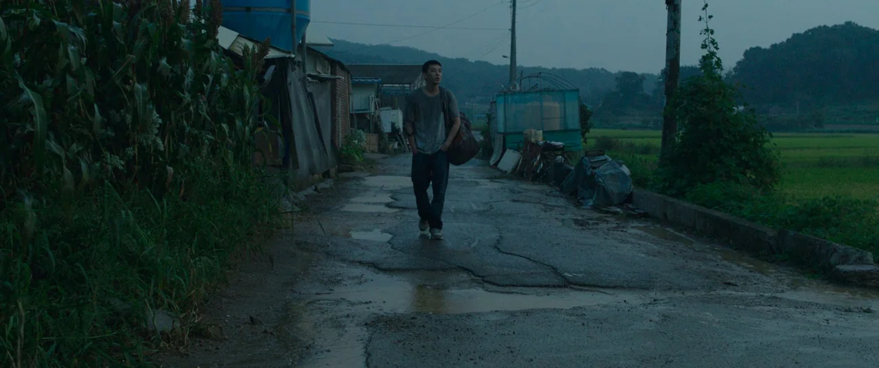 Burning (2018) movie still: fullbody — A wet asphalt road runs through a rural area lined with corn stalks. A young man walks toward the camera with…