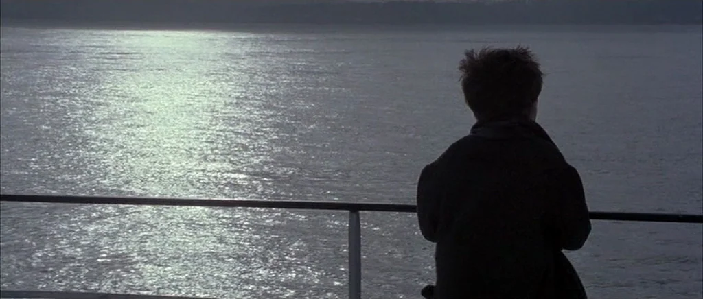 Jude (1996) movie still: medium — A dark metal railing separates the viewer from a large expanse of rippling water. A young person viewed from…