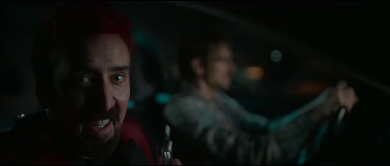 Sympathy For The Devil (2023) movie still: closeup — The interior of a car at night with a man in the foreground. He looks at the camera while the…