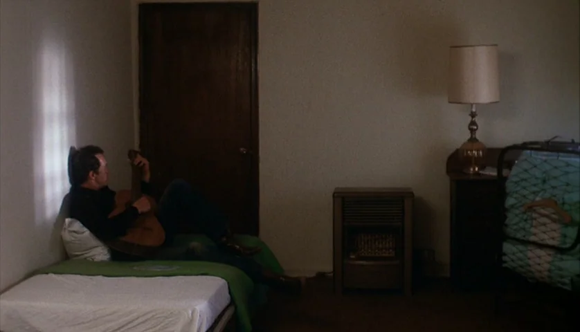 Cockfighter (1974) movie still: medium — A sparsely furnished motel room features white walls, a single door, and two beds. A man lies on a green…