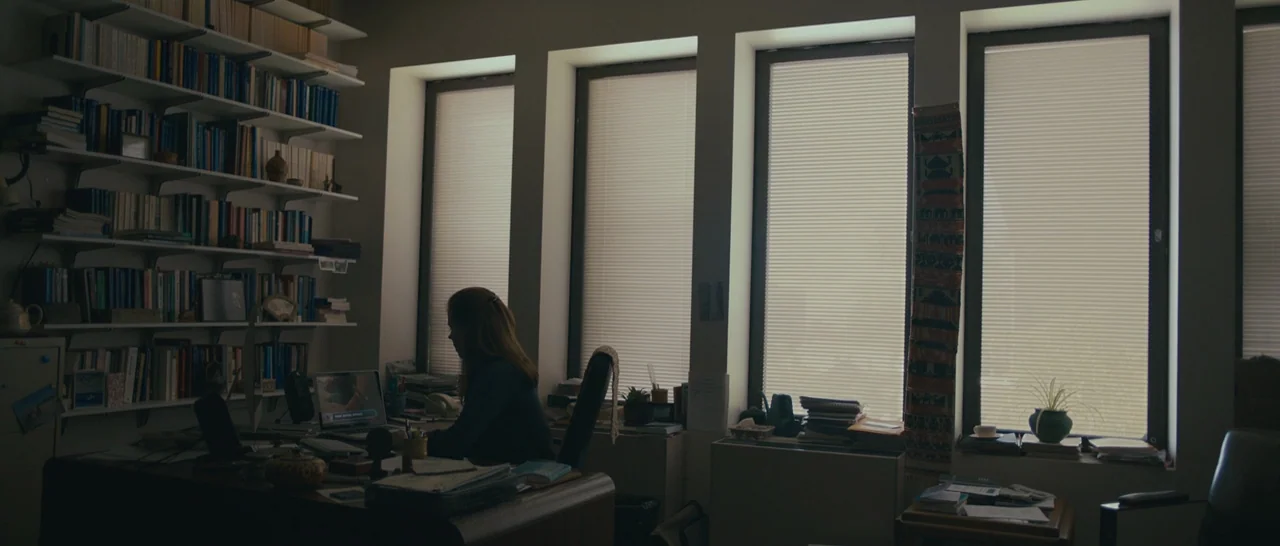 Arrival (2016) movie still: wide — A quiet home office features a wall of bookshelves and large windows with blinds. A woman sits at a wooden desk…