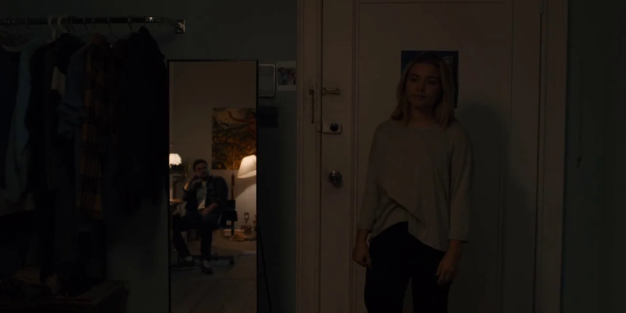Midsommar (2019) movie still: medium — A dimly lit apartment foyer features a tall mirror reflecting a seated man nearby. The woman faces forward in…