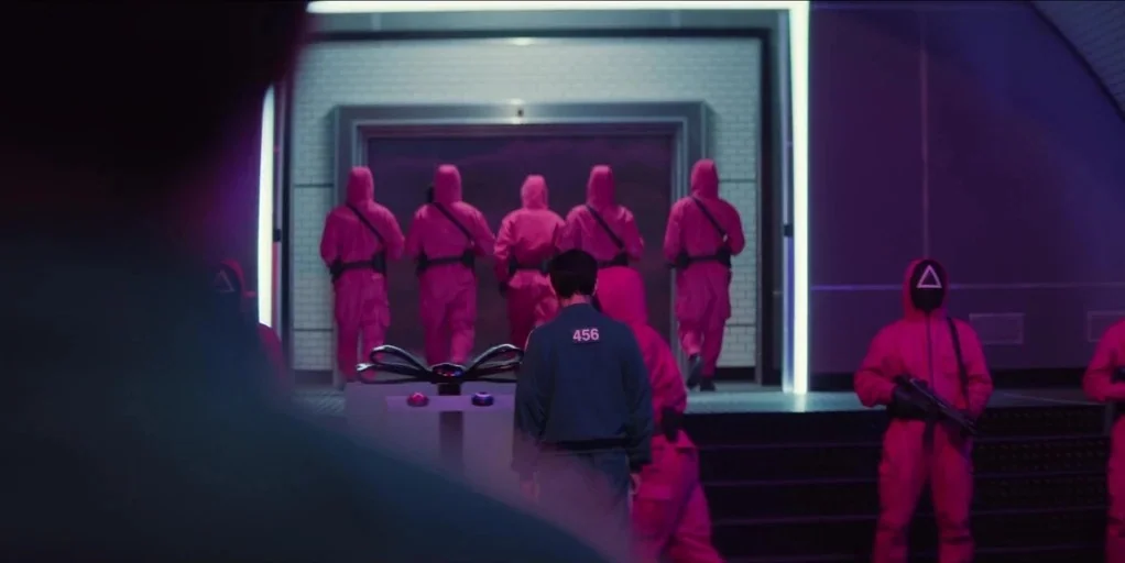 Squid Game (2024) movie still: medium — A dark, futuristic-looking hallway features white tiled walls and a large, bright doorway ahead. Several…