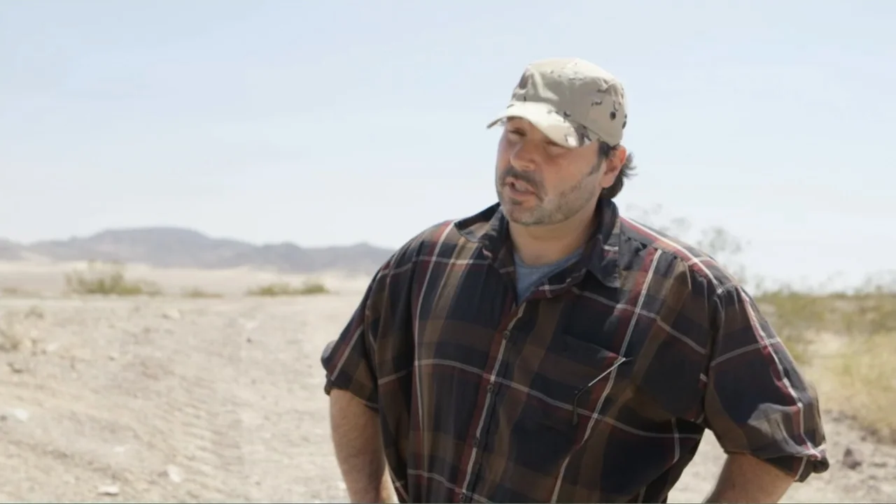 Savageland (2015) movie still: medium — A dry desert landscape with sparse vegetation stretches out under a clear sky. A man wearing a plaid shirt…