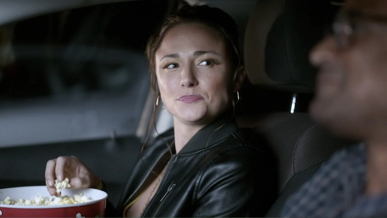 Ask Me to Dance (2022) movie still: medium — A dark car interior at night with a person in the foreground blurred. A woman in a leather jacket holds…