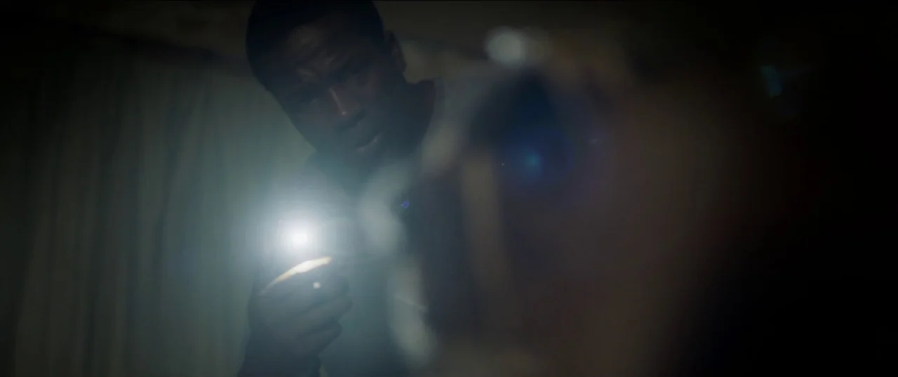His House (2020) movie still: medium — A dark, dimly lit interior space with shadows obscuring the surroundings. A man shines a bright flashlight…