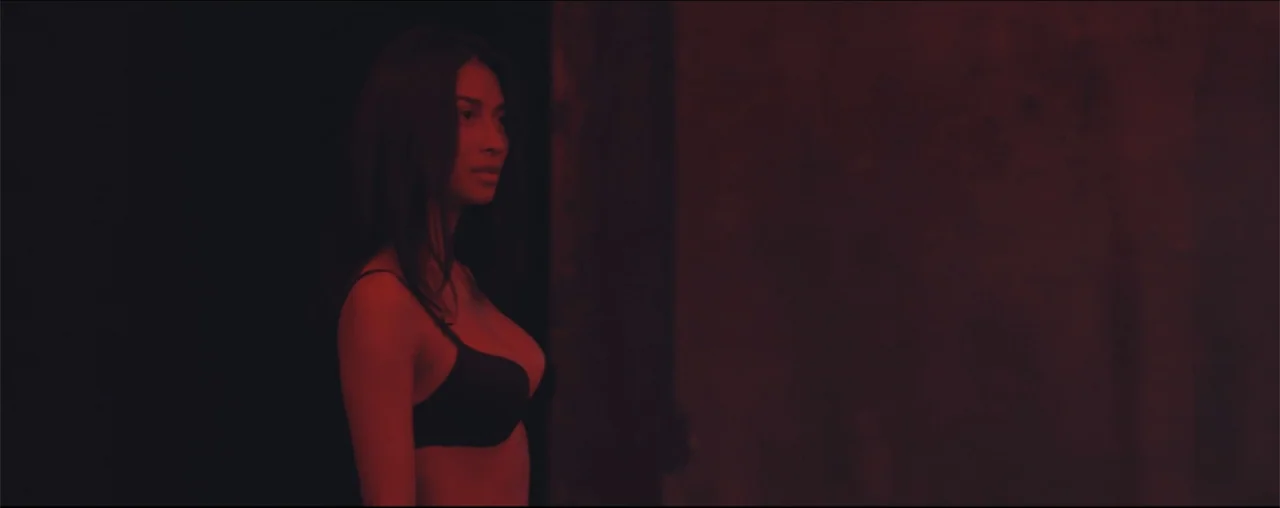 El Último Juego (2025) movie still: medium — A dark, minimally furnished interior space is bathed in intense red light. A young woman in a black…