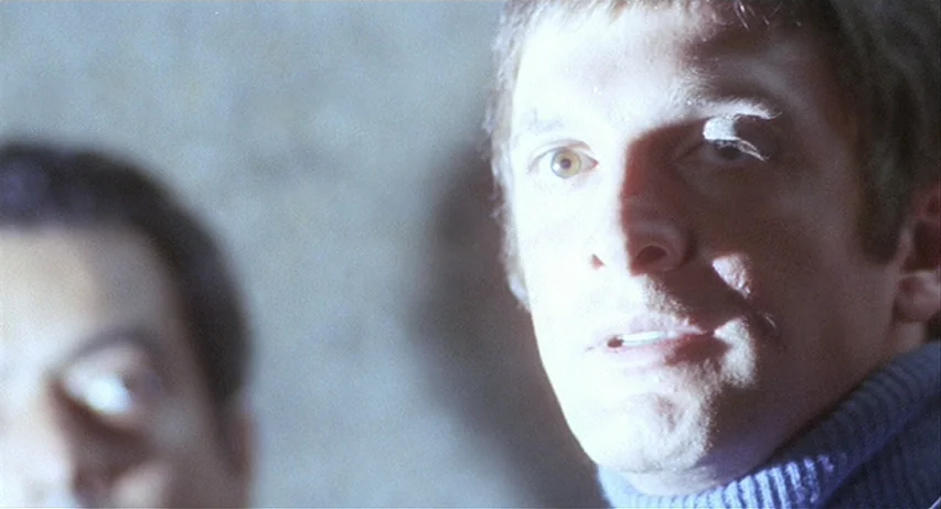 The Fifth Cord (1971) movie still: closeup — A dark, textured interior space provides a background for two men. One man in a blue turtleneck looks…
