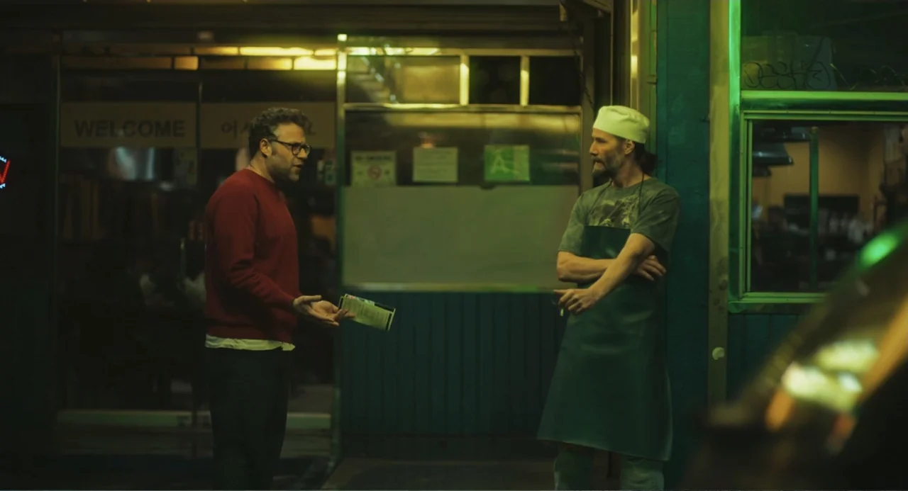 Good Fortune (2025) movie still: medium — A nighttime exterior scene outside a commercial building with green neon lighting. One man speaks to…