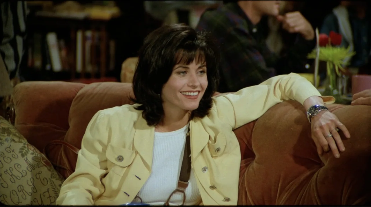 Friends: The Reunion (2021) movie still: medium — A woman sits on a plush velvet couch inside a cozy decorated cafe. The woman leans back against…