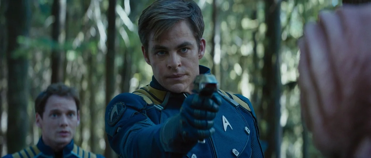 Star Trek Beyond (2016) movie still: medium — A dense patch of woodland with tall tree trunks provides a shadowed backdrop. One man holds a phaser…