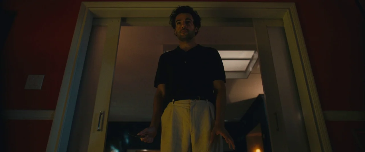 Sanctuary (2023) movie still: medium — A man stands centered in a doorway against a dark, red-walled background. He holds a small metallic object in…
