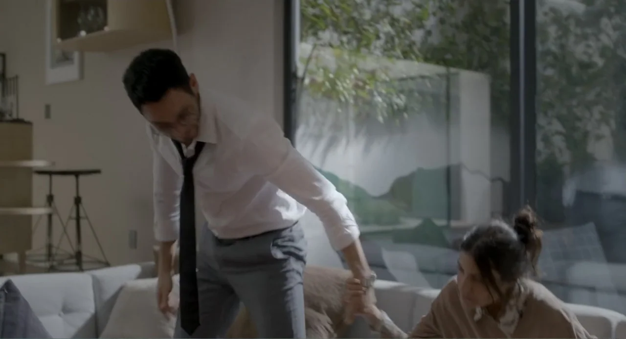 Straight (2024) movie still: medium — A modern interior with a sofa and a large floor-to-ceiling glass window behind it. A man stands over a seated…