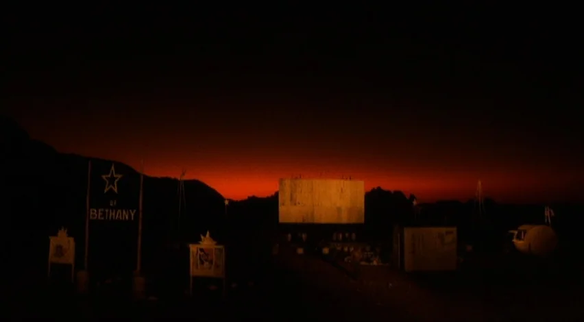Dust Devil (1992) movie still: establishing — A dark drive-in theater sits against a deep red sunset in a mountainous region. The wooden screen…