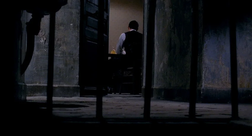 The Man Who Cried (2000) movie still: medium — A dark, spartan room with iron bars in the foreground and gray walls. A man in a dark vest sits alone…