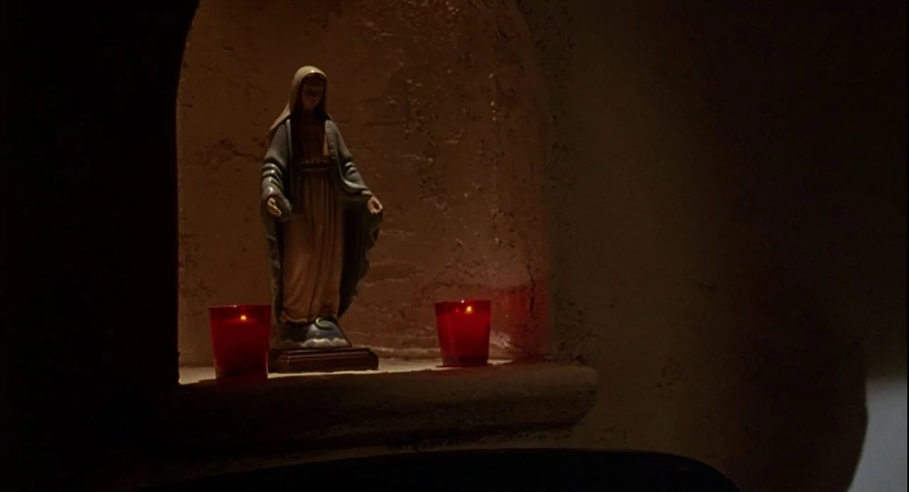 Psycho III (1986) movie still: medium — A small carved wall niche contains a miniature statue of the Virgin Mary. Two red glass votive candles sit…