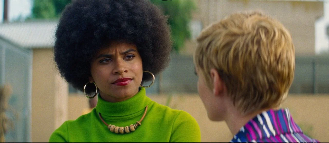 Seberg (2019) movie still: medium — A bright suburban outdoor space features soft sunlight hitting the two subjects. A woman with an afro in a green…