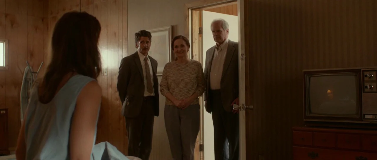 Faults (2014) movie still: medium — A wood-paneled room contains an old wooden television console on a dresser. Three people stand inside an open…