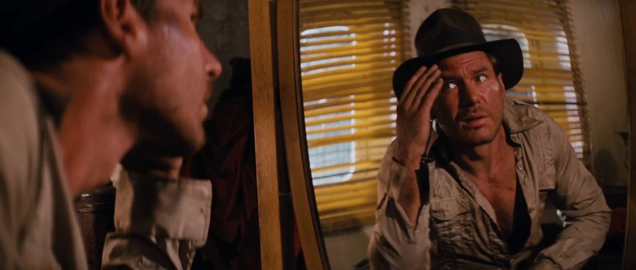 Raiders Of The Lost Ark (1981) movie still: medium — A man in a fedora examines his reflection in a wooden-framed mirror. Sunlight streams through…