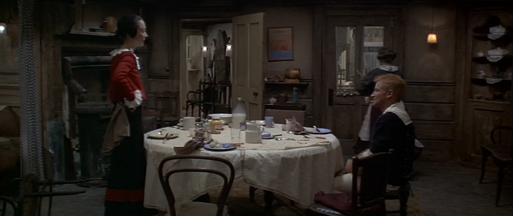 Popeye (1980) movie still: wide — A rustic dining room features wooden walls, a round table with a white cloth. A woman stands near the table while…