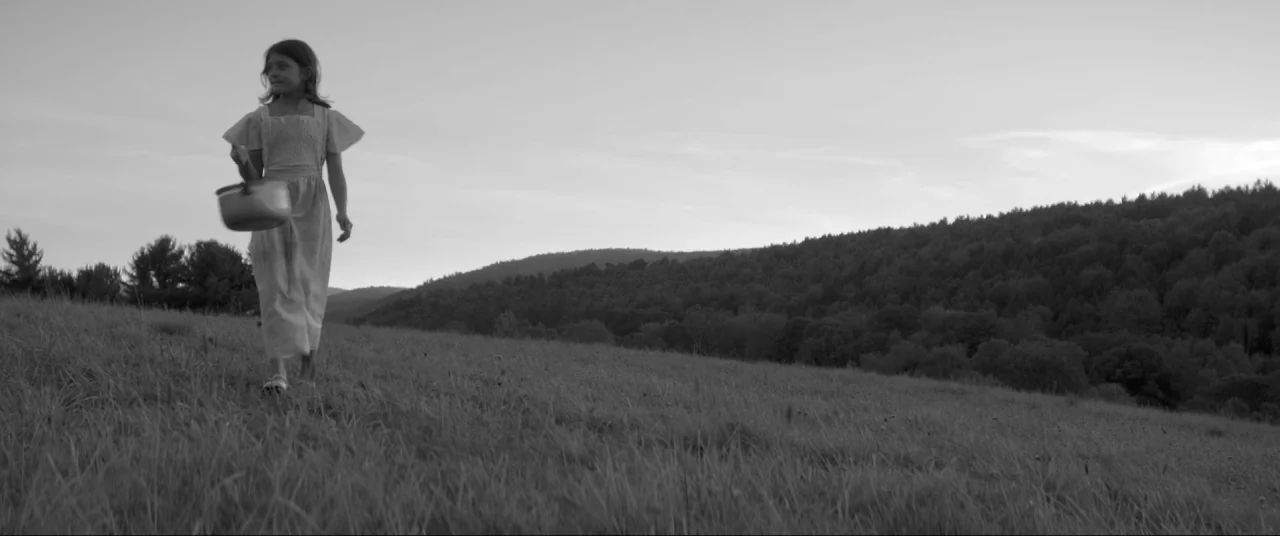 The Eyes of My Mother (2016) movie still: fullbody — An open grassy meadow slopes upward toward a dense treeline against the sky. A young girl in a…