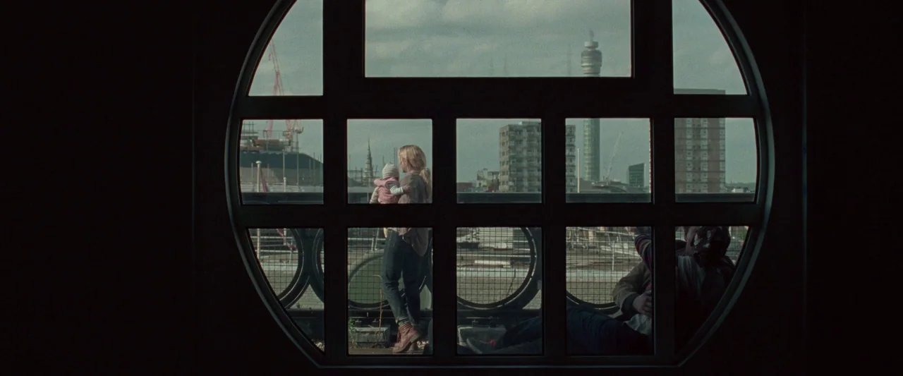 The End We Start From (2023) movie still: wide — A view through a circular window shows a woman holding a baby on an urban balcony. Beyond the metal…
