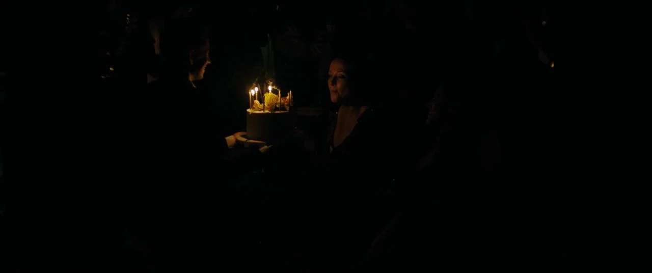Saint Maud (2019) movie still: medium — A dimly lit room with heavy shadows surrounding a single point of bright light. Two people sit side by side…