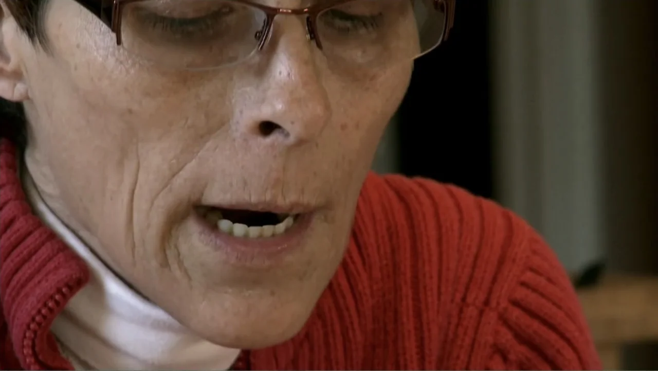 Wartorn: 1861-2010 (2010) movie still: closeup — A close-up view of a woman indoors in a dimly lit space. She wears glasses and a red sweater while…