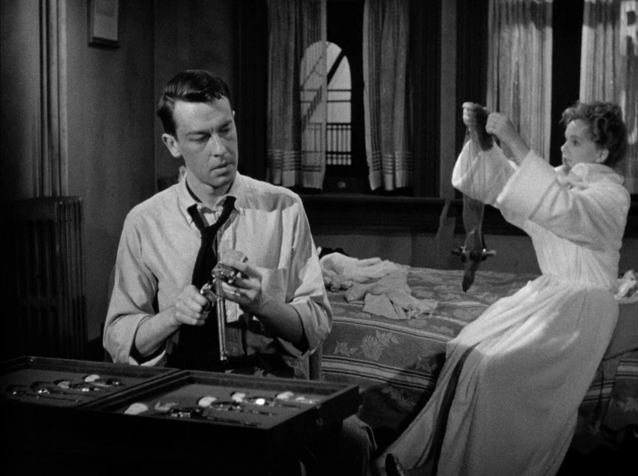 Gun Crazy (1950) movie still: medium — A dimly lit room contains a bed, a window with curtains, and a radiator. The man holds a revolver from a…