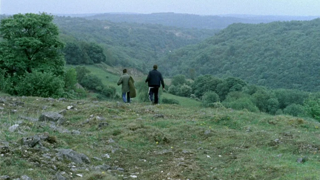 Dead Man’s Shoes (2004) movie still: wide — A grassy, rocky hillside overlooks a sprawling expanse of rolling, forested hills. Two men walk away…