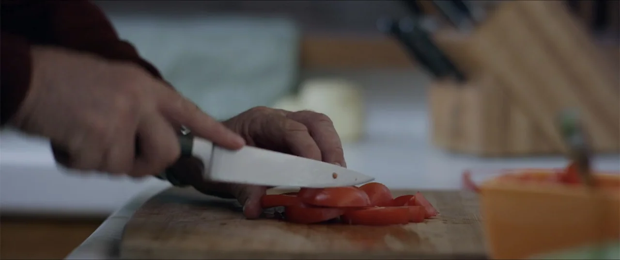 Captive (2020) movie still: closeup — A wooden cutting board rests on a white kitchen surface with a knife. A person is slicing a red tomato into…