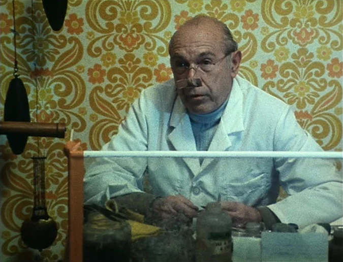 Daguerréotypes (1976) movie still: medium — An older man sits at a workspace with vintage-patterned wallpaper in the background. He wears a lab coat…