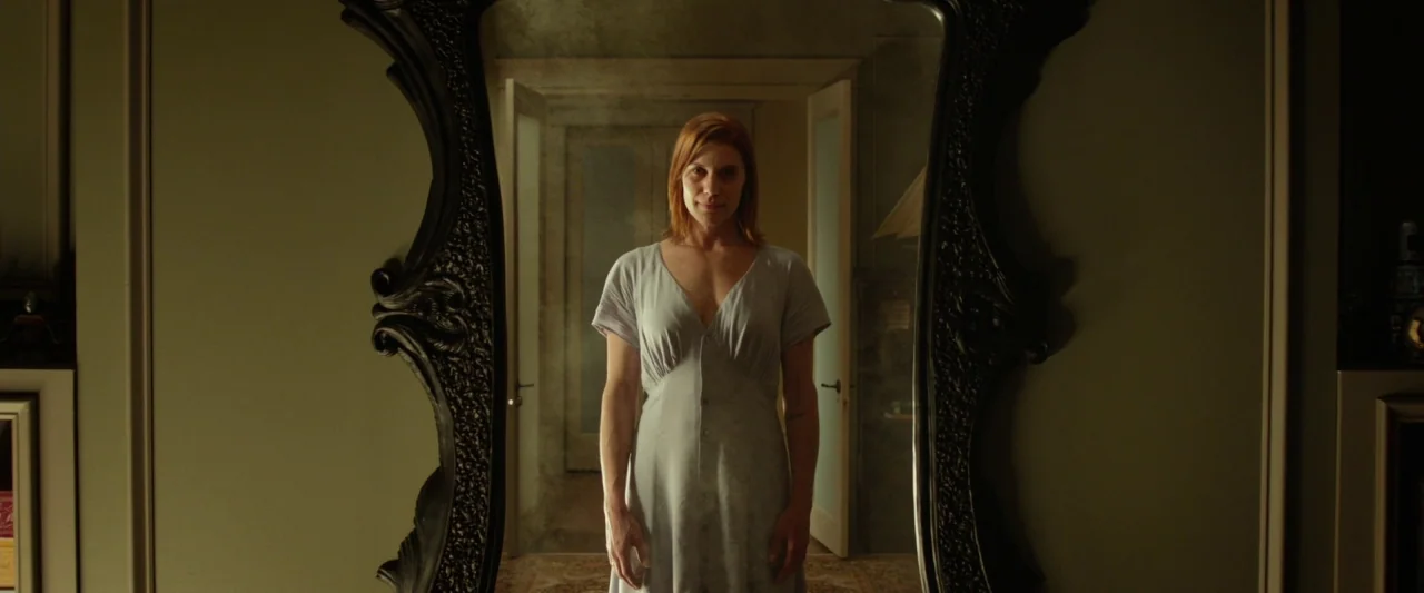 Oculus (2013) movie still: medium — A woman stands centered inside a large, ornate black-framed mirror reflecting a lit hallway. The woman is…