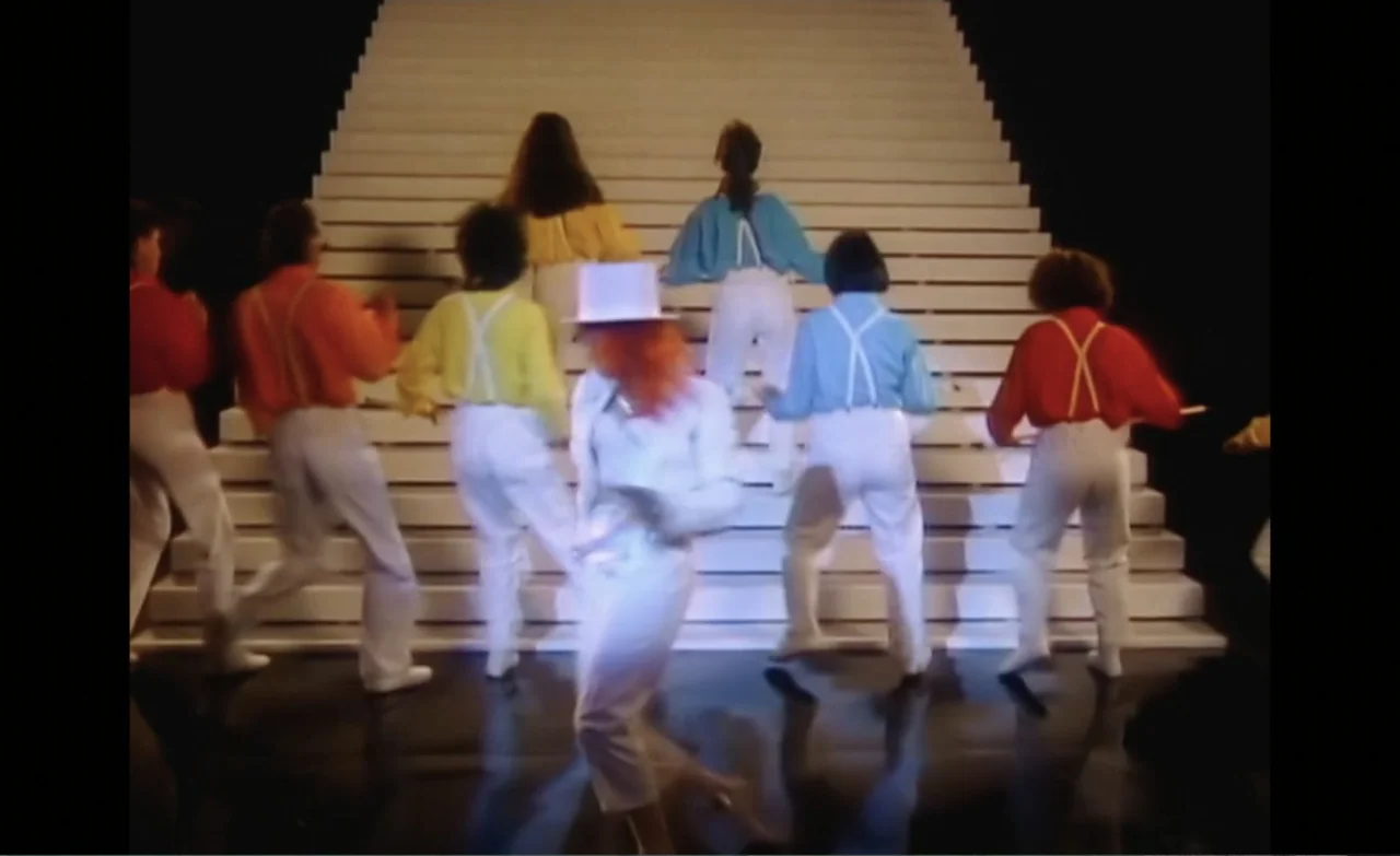 Let the Canary Sing (2023) movie still: fullbody — A group of dancers performs on a brightly lit white staircase set. The performers move upward…
