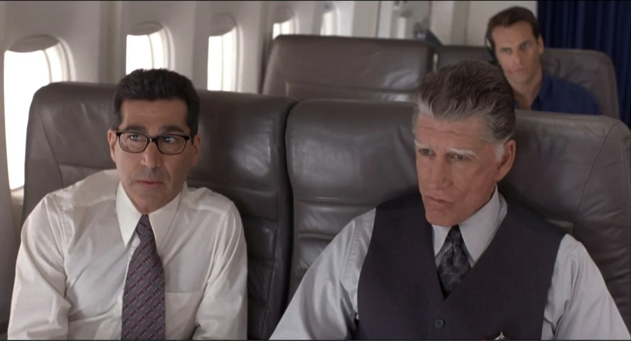 Air Rage (2001) movie still: medium — Two men sit side by side in gray leather seats inside an airplane cabin. The men stare ahead while a third man…